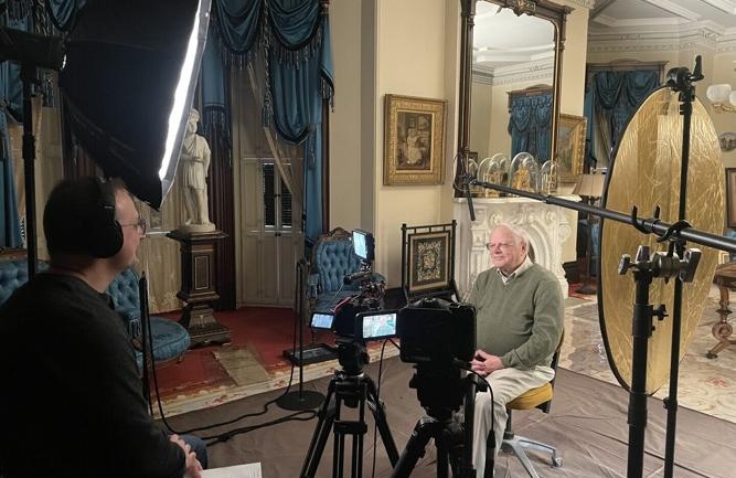 Shane Johnson, wearing a headset with a camera, interviews George DeMass. They are seated in the historic parlor.